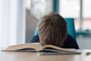 Pessoa com cabelo castanho claro apoia a cabeça sobre um livro aberto em uma mesa, em ambiente interno com luz natural.