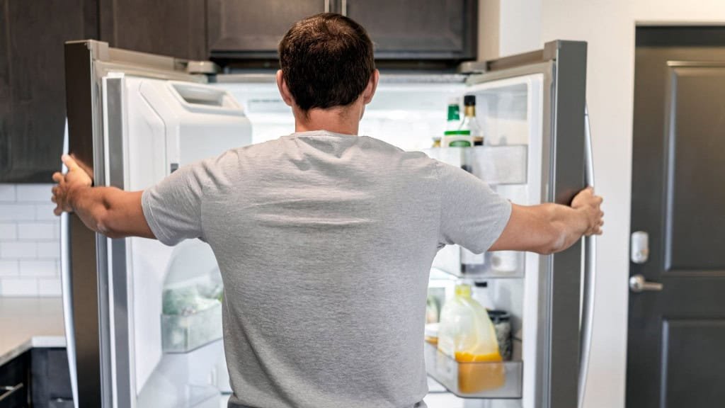 🍽️ Costas de um homem faminto abrindo as portas da geladeira, eletrodoméstico moderno, procurando comida entre condimentos e suco em uma cozinha contemporânea.