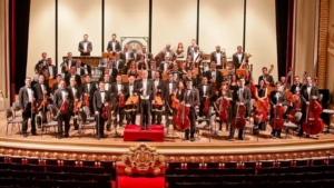 Orquestra sinfônica reunida no palco de um teatro histórico, com músicos e maestro em formação clássica, simbolizando a força da música como expressão do Espírito do Natal e da comunhão humana.