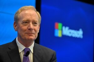 Um homem com cabelo claro e uma expressão serena está em um evento, usando um paletó escuro e uma gravata roxa. Ao fundo, há um logotipo da Microsoft em cores azul, verde, amarelo e vermelho, em um painel azul.