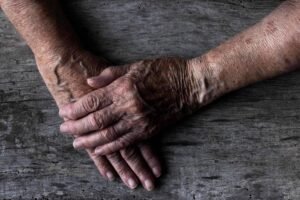 Mãos de pessoa idosa cruzadas repousam sobre uma mesa de madeira rústica. A pele apresenta rugas, manchas e textura característica do envelhecimento.