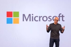 Executivo da Microsoft em pé à direita, gesticulando com as mãos durante apresentação. Ao fundo, logo colorido da Microsoft em fundo branco.