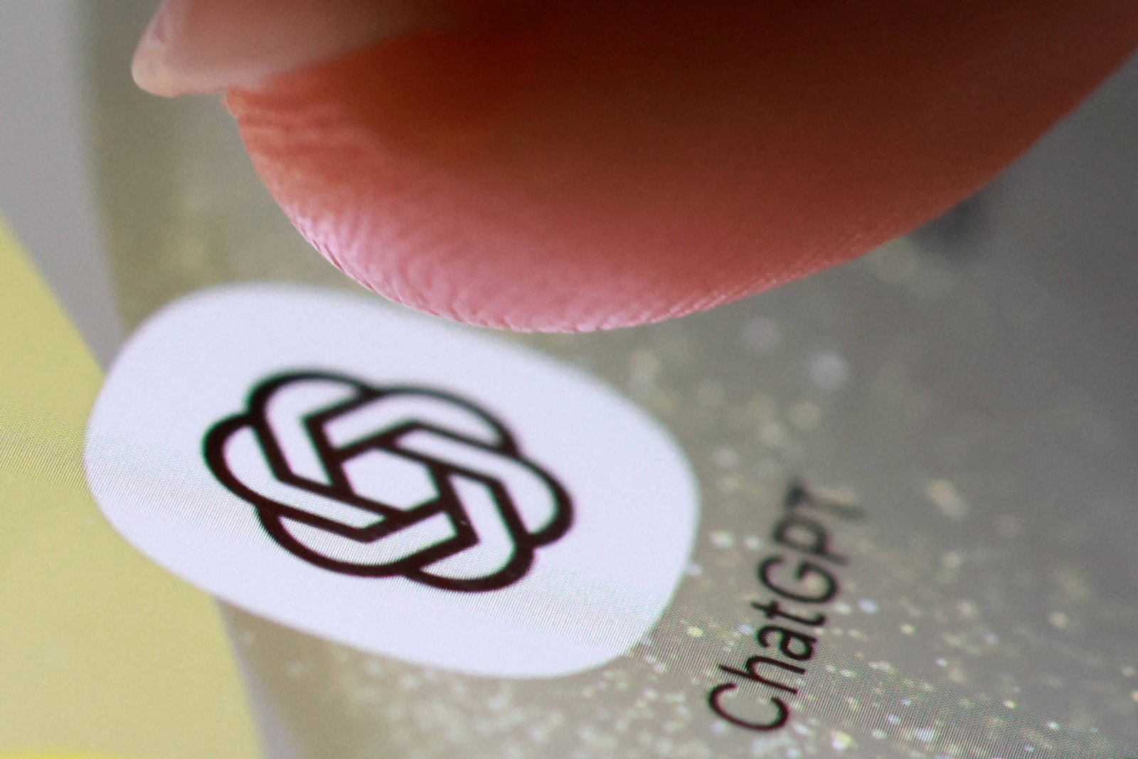 Dedo indicador próximo ao ícone do aplicativo ChatGPT exibido na tela de um smartphone. O ícone é branco com o logotipo preto entrelaçado e o texto
