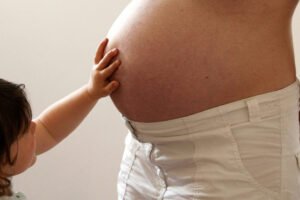 Criança com cabelo castanho toca com a mão a barriga de mulher grávida vestindo calça branca. Fundo neutro claro.