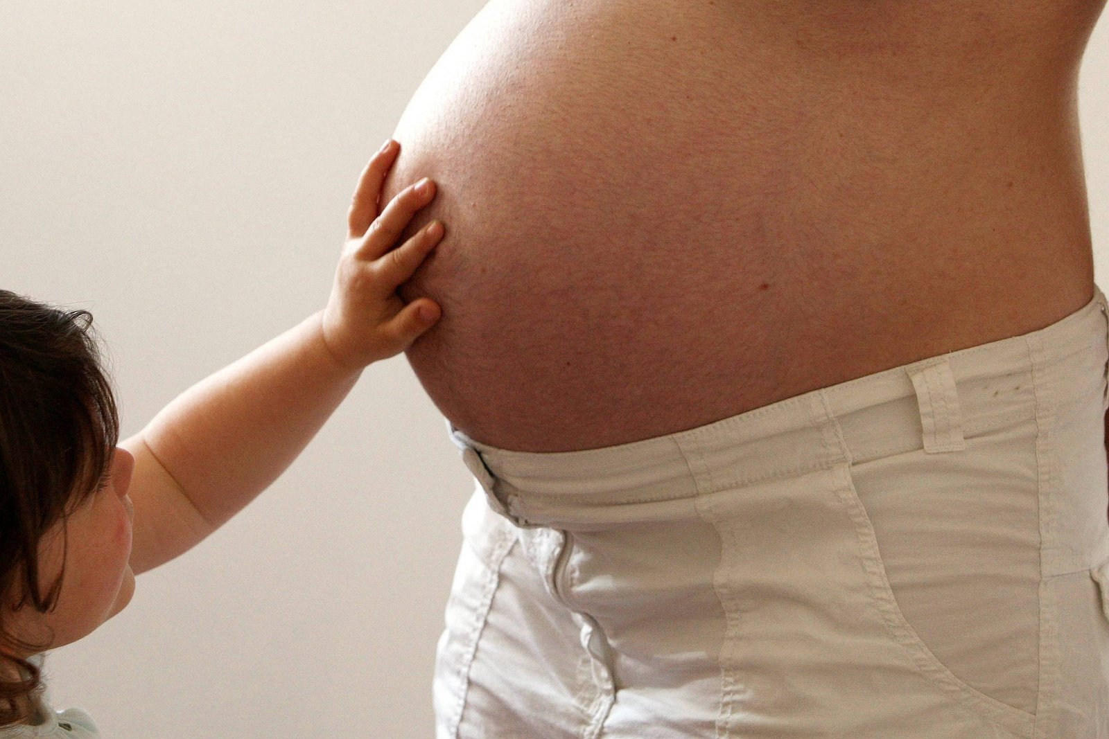 Criança com cabelo castanho toca com a mão a barriga de mulher grávida vestindo calça branca. Fundo neutro claro.