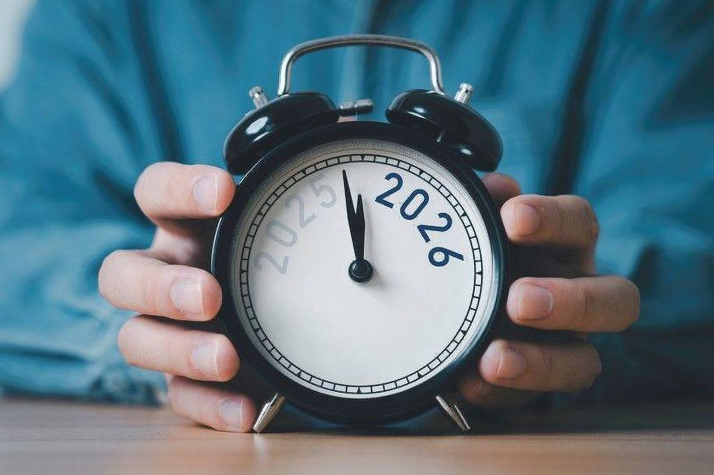 Relógio despertador preto com mostrador branco segurado por duas mãos. No lugar dos números tradicionais, aparece o ano 2026 em destaque azul, com o ponteiro marcando meia-noite.