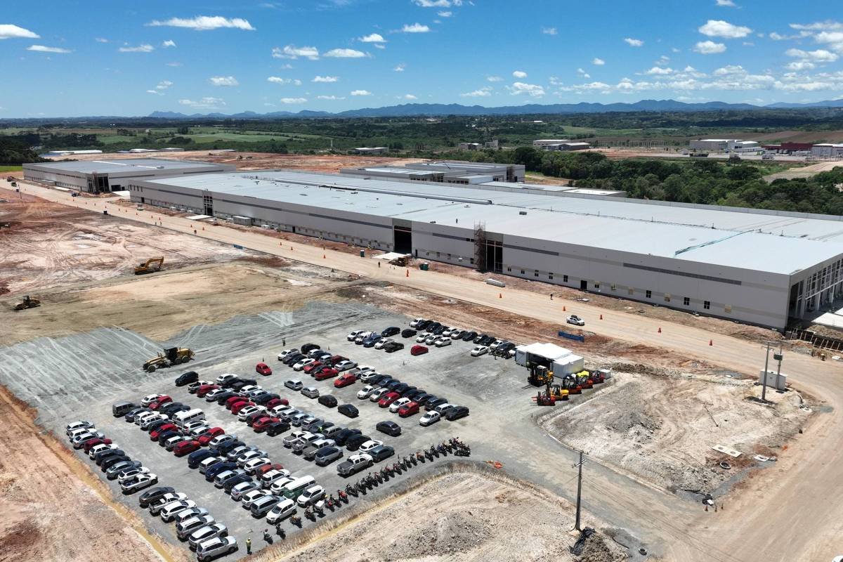 Galpão industrial extenso em construção em área aberta, com estacionamento organizado de veículos ao lado. Terreno ao redor está em preparação, com máquinas e terra exposta. Céu claro com poucas nuvens e montanhas ao fundo.