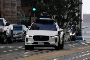 Carro branco autônomo equipado com sensores no teto circula em via urbana com semáforo verde. Veículos e motociclista aparecem ao fundo em ambiente de cidade.