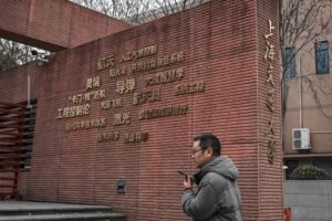Homem de casaco cinza caminha segurando celular em frente a muro de tijolos vermelhos com texto em chinês dourado. Ambiente urbano com árvores sem folhas ao fundo.