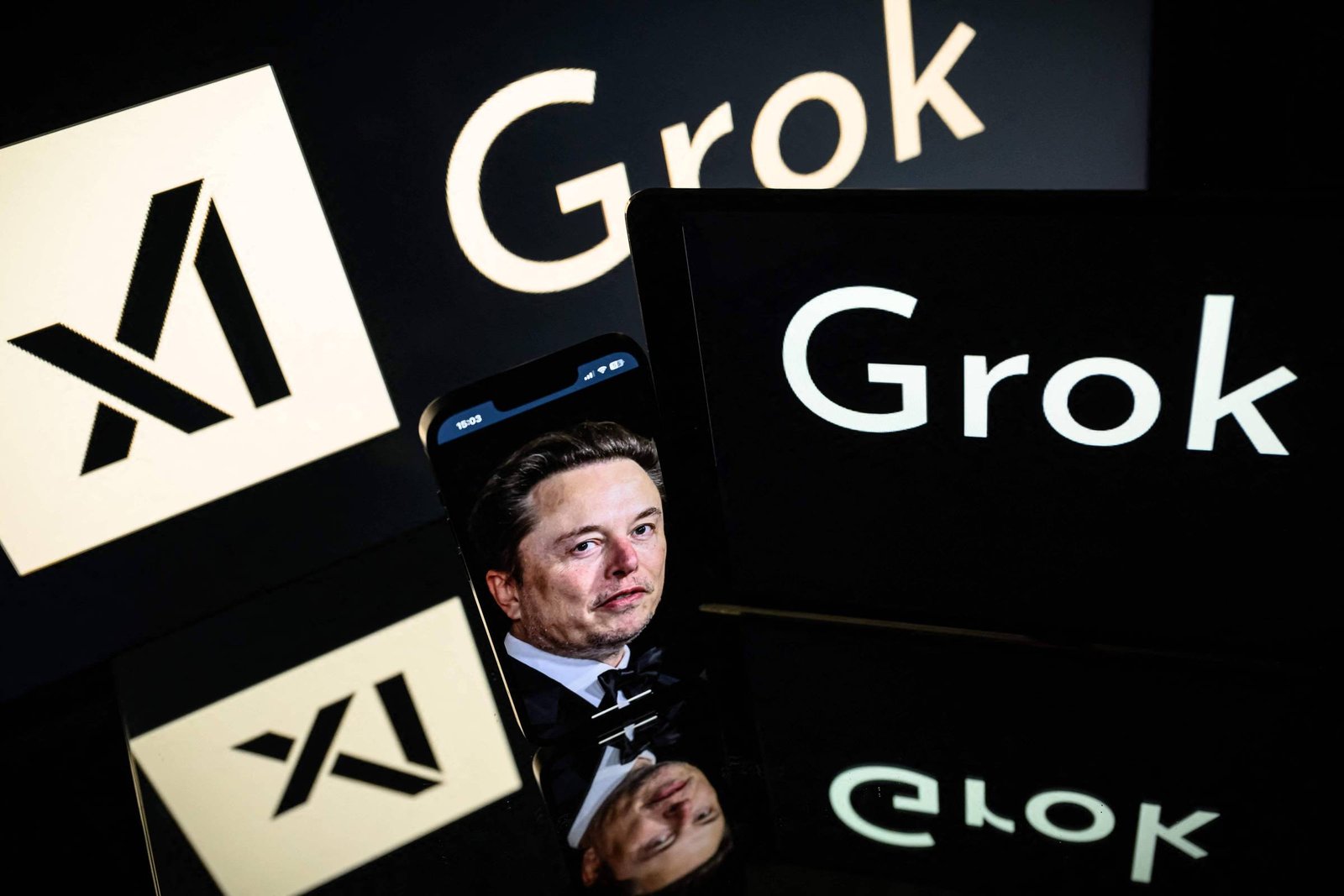 Tela de computador e smartphone exibem os logos do Grok e X. Reflexo do rosto de Elon Musk aparece na tela do smartphone em primeiro plano.