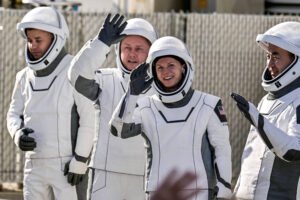 Quatro astronautas vestindo trajes espaciais brancos com detalhes cinza estão alinhados em ambiente externo. Dois deles acenam para a câmera, enquanto os outros observam ao redor. Um deles exibe a bandeira dos Estados Unidos na manga.
