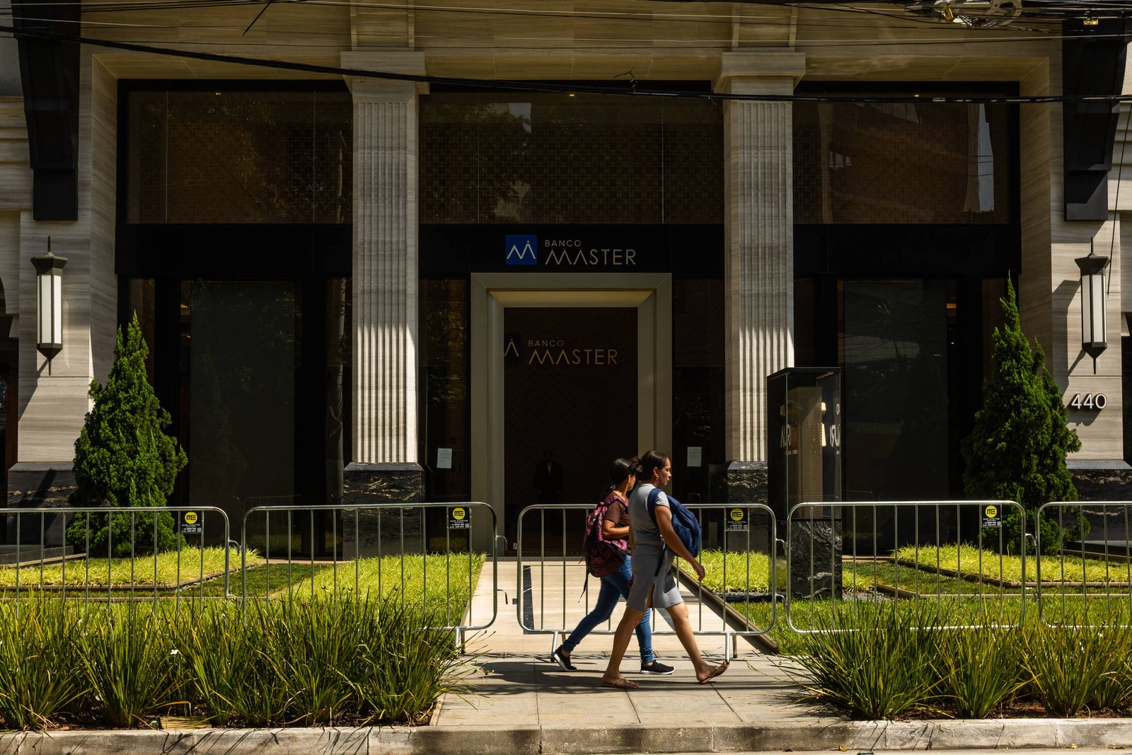 Duas pessoas caminham lado a lado na calçada em frente a um prédio com fachada clássica, com colunas e vegetação aparada. O prédio tem uma placa com o nome