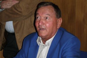 Homem de meia-idade veste paletó azul e camisa branca, sentado à mesa de madeira, segurando caneta e olhando para frente. Fundo com parede de madeira e outra pessoa parcialmente visível ao lado.