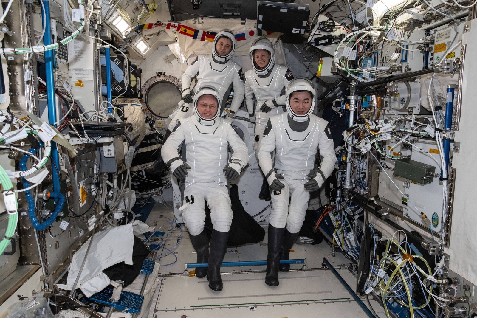 Quatro astronautas vestindo trajes espaciais brancos posam no interior da Estação Espacial Internacional, cercados por equipamentos, cabos e painéis técnicos. Dois estão agachados à frente e dois estão em pé atrás, em ambiente confinado e tecnológico.
