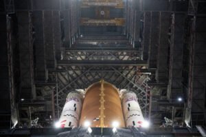 Tanque externo laranja e dois foguetes laterais brancos de foguete vistos de baixo para cima dentro de hangar industrial com estrutura metálica e iluminação artificial.