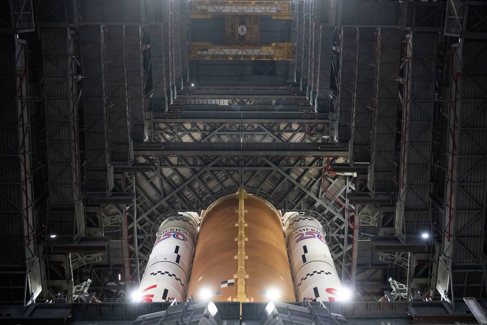 Tanque externo laranja e dois foguetes laterais brancos de foguete vistos de baixo para cima dentro de hangar industrial com estrutura metálica e iluminação artificial.