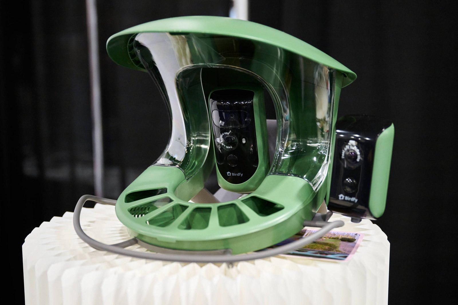 Dispositivo tecnológico verde com design curvo e câmeras embutidas, exibido sobre superfície branca texturizada. Fundo escuro destaca o aparelho.