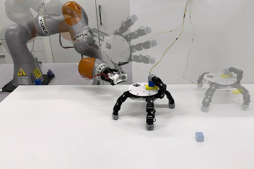Braço robótico cinza e laranja manipula um pequeno cubo azul sobre mesa branca. Robô de três pernas está na mesa, com imagem fantasma indicando movimento para a direita. Fundo branco com paredes e armários.