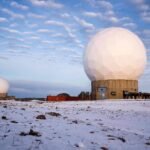 Três grandes radares esféricos brancos sobre bases cilíndricas em terreno coberto por neve. Céu azul com nuvens dispersas ao fundo.