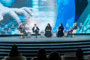 Cinco pessoas sentadas em cadeiras no palco de um evento, com grande tela digital ao fundo exibindo imagem de mãos digitando em teclado. Público assiste em poltronas na frente do palco, sob iluminação azulada e lustre grande no teto.