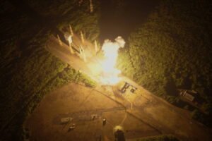 Foguete é lançado de plataforma iluminada à noite, com chama intensa e fumaça visível. Área ao redor é coberta por vegetação densa e algumas estruturas próximas.