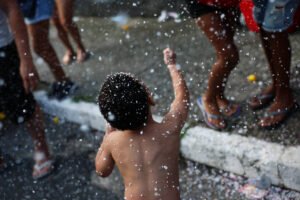A imagem mostra uma criança pequena, sem camisa, de costas, levantando o braço em um ambiente festivo. Ao redor, há várias pessoas, algumas com pernas visíveis, e o chão está coberto de espuma. A cena transmite um clima de alegria e celebração.