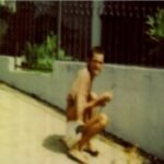 Homem sem camisa e de bermuda branca agachado ao lado de um muro branco em área externa. Ao fundo, grades pretas e plantas verdes.