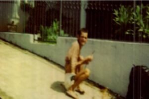 Homem sem camisa e de bermuda branca agachado ao lado de um muro branco em área externa. Ao fundo, grades pretas e plantas verdes.