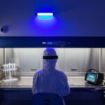 Pessoa vestida com traje de proteção completo sentada diante de uma cabine de fluxo laminar iluminada, com luz azul acesa acima. Equipamentos de laboratório, incluindo suporte para tubos e dispositivo eletrônico, estão posicionados dentro da cabine.