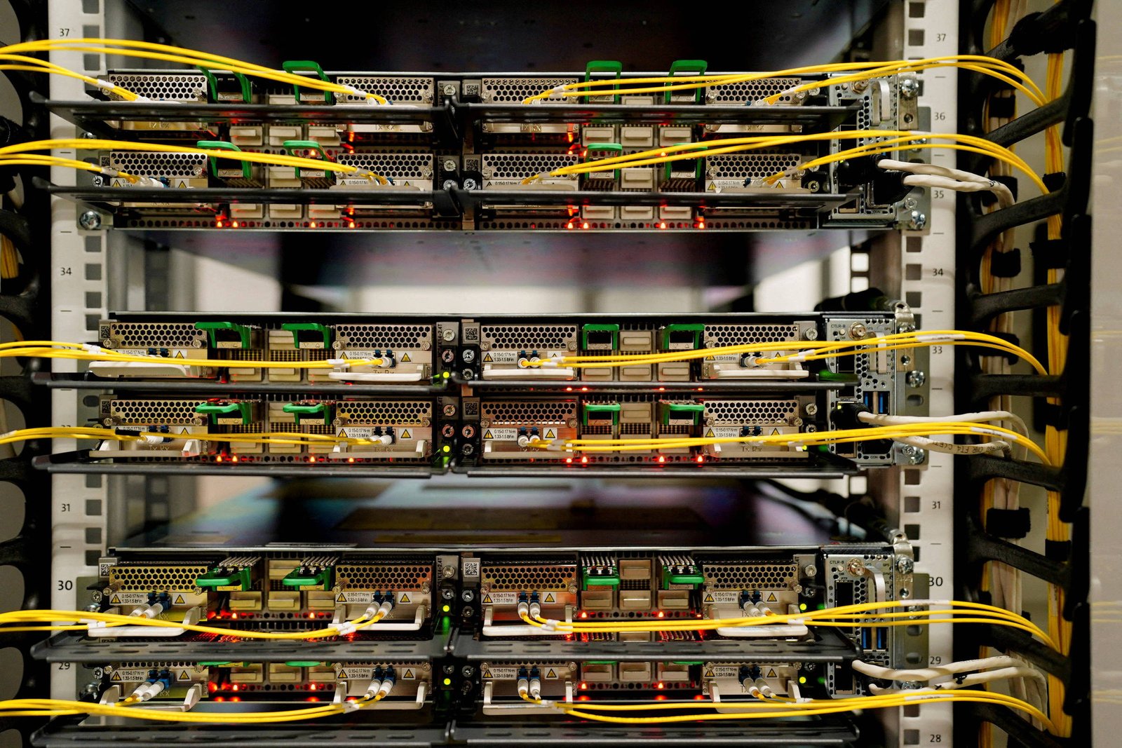 Rack de servidores com múltiplos módulos de hardware conectados por cabos amarelos de fibra óptica. Luzes indicadoras vermelhas e verdes estão acesas nos equipamentos, indicando atividade. O ambiente é interno, com estrutura metálica e organização técnica visível.