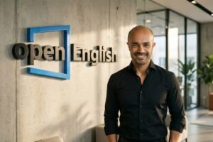 Homem calvo, de pele clara, veste camisa preta e sorri em pé ao lado de parede cinza com logo 3D azul e preto da Open English. Ambiente interno moderno com janelas grandes, plantas e móveis ao fundo.