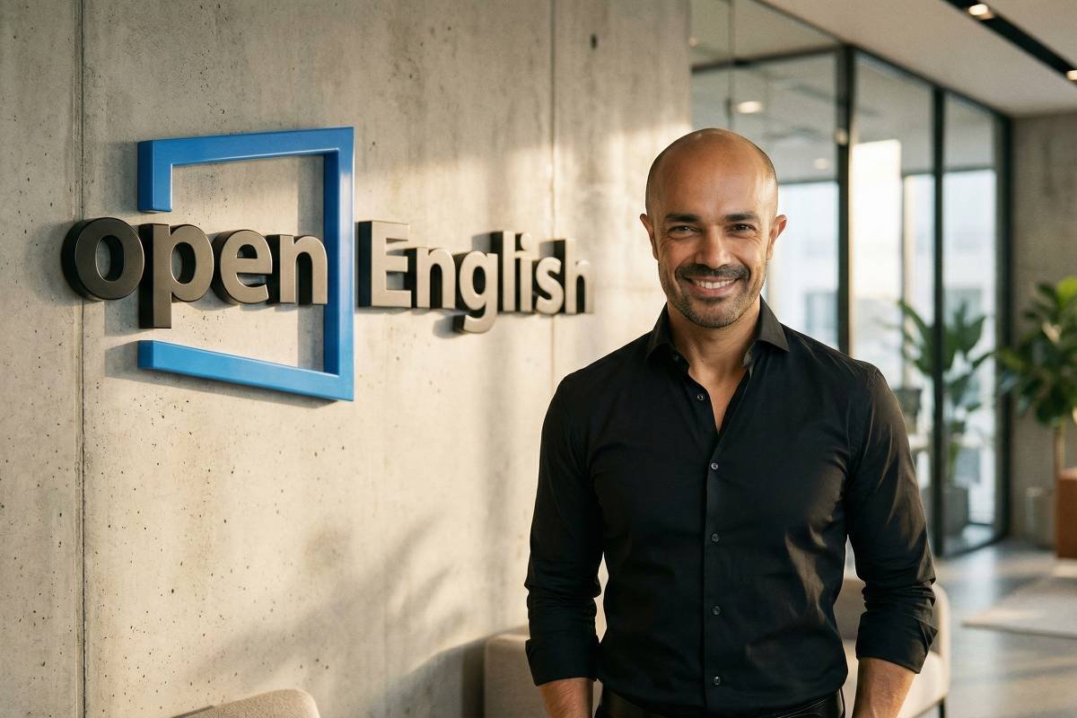 Homem calvo, de pele clara, veste camisa preta e sorri em pé ao lado de parede cinza com logo 3D azul e preto da Open English. Ambiente interno moderno com janelas grandes, plantas e móveis ao fundo.