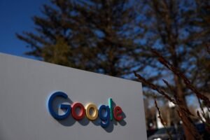 Logotipo do Google em letras coloridas fixada em placa branca ao ar livre, com árvores e céu azul ao fundo.