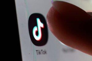Dedo indicador próximo ao ícone do aplicativo TikTok exibido em tela de smartphone. Ícone apresenta símbolo musical estilizado em cores neon sobre fundo preto.