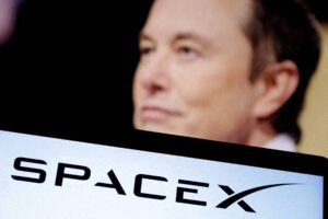 Elon Musk aparece desfocado ao fundo, com foco em tela à frente exibindo o logo da SpaceX em preto sobre fundo claro.