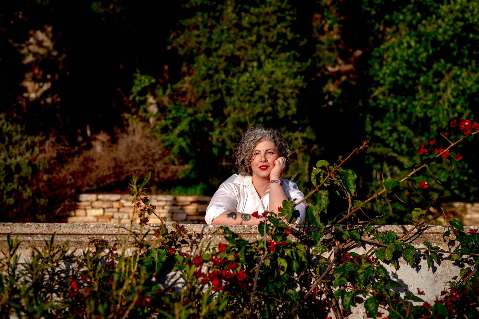 Mulher de cabelo grisalho encaracolado e batom vermelho apoia o rosto com a mão direita sobre um muro baixo em jardim com folhagens e flores vermelhas. Fundo com árvores e arbustos em luz natural.