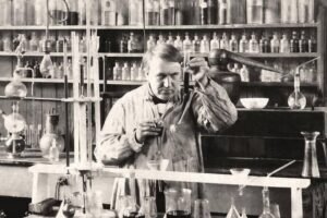 Homem com jaleco manipula frasco em laboratório antigo com prateleiras cheias de frascos e equipamentos de vidro ao fundo.