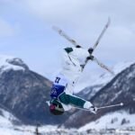 Esquiadora com uniforme branco e verde executa salto mortal invertido com esquis cruzados em área nevada de montanha. Céu nublado e picos cobertos de neve ao fundo.