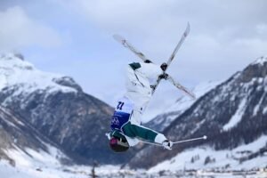 Esquiadora com uniforme branco e verde executa salto mortal invertido com esquis cruzados em área nevada de montanha. Céu nublado e picos cobertos de neve ao fundo.