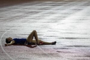 Atleta vestindo uniforme azul e tênis vermelhos está deitada de costas na pista de corrida molhada, com um braço estendido e uma perna dobrada, aparentemente descansando ou exausta.