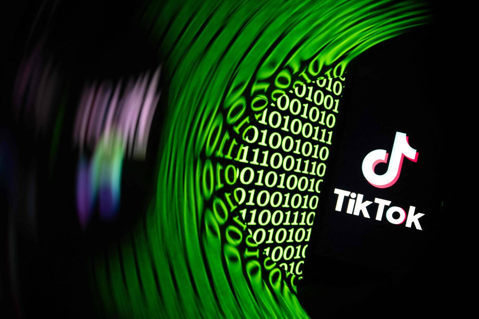 Tela de celular exibe o logo do TikTok em fundo preto, ao lado de uma sequência de números binários verdes (0 e 1) em padrão curvo sobre fundo escuro.