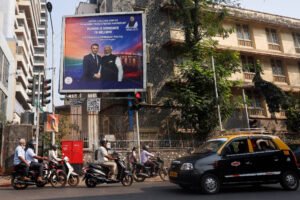 Motociclistas e um táxi aguardam no semáforo em rua movimentada com edifícios ao fundo. Outdoor exibe mensagem de boas-vindas a líder indiano, com foto dele e de outra pessoa.