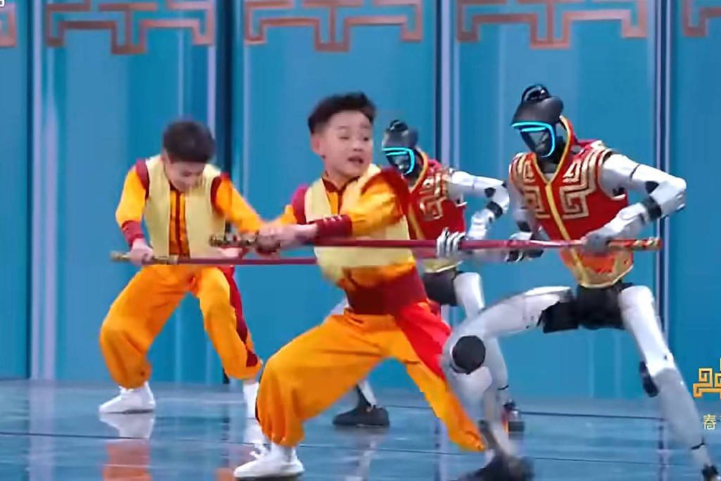 Duas crianças vestidas com roupas amarelas e laranja seguram bastões enquanto dançam ao lado de dois robôs com trajes vermelhos e detalhes dourados em palco azul.