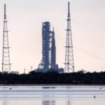 Foguete posicionado em plataforma de lançamento cercada por quatro torres altas com cabos de sustentação. Vegetação baixa e corpo d