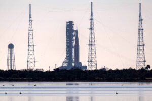 Foguete posicionado em plataforma de lançamento cercada por quatro torres altas com cabos de sustentação. Vegetação baixa e corpo d