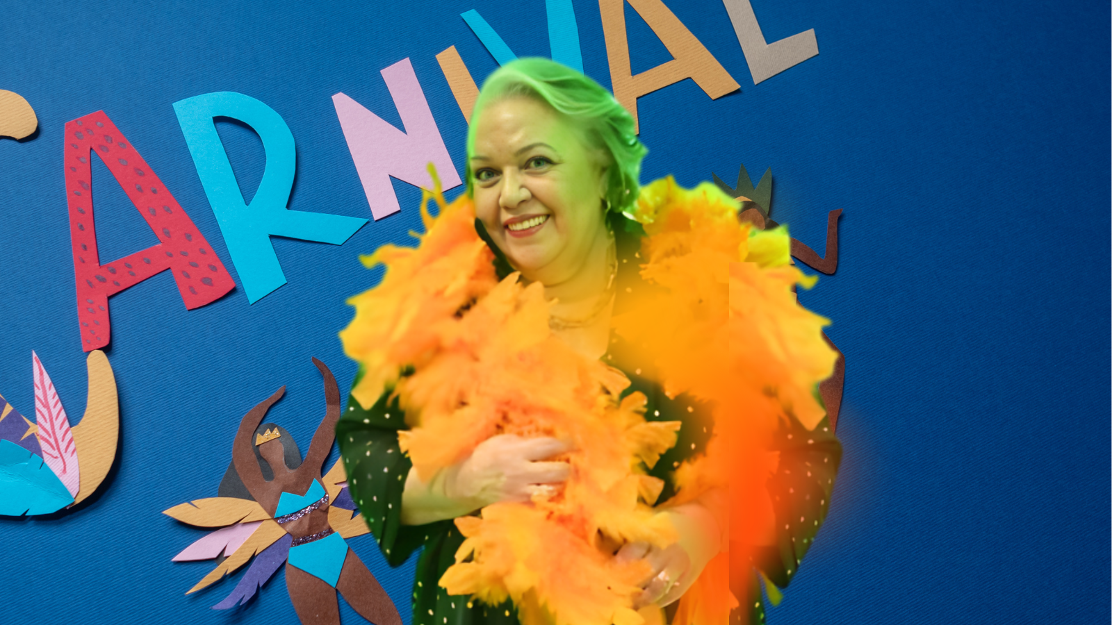 Myrinha Vasconcellos sorrindo com plumas amarelas em frente a painel azul decorado com letras coloridas formando a palavra “Carnaval”.