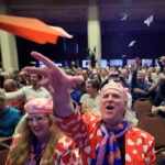 A imagem mostra uma grande audiência em um evento, com várias pessoas sentadas em cadeiras. No primeiro plano, um homem com um paletó colorido com corações e uma gravata laranja está lançando um avião de papel. Ao lado dele, uma mulher com um chapéu rosa e uma blusa azul observa. Ao fundo, outras pessoas também estão lançando aviões de papel, criando um ambiente festivo e animado.