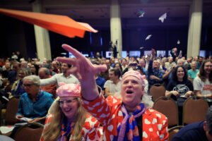 A imagem mostra uma grande audiência em um evento, com várias pessoas sentadas em cadeiras. No primeiro plano, um homem com um paletó colorido com corações e uma gravata laranja está lançando um avião de papel. Ao lado dele, uma mulher com um chapéu rosa e uma blusa azul observa. Ao fundo, outras pessoas também estão lançando aviões de papel, criando um ambiente festivo e animado.