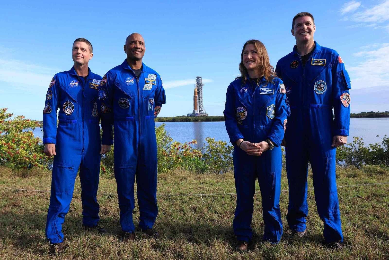 Quatro astronautas em trajes azuis posam em pé sobre grama com vegetação e corpo d