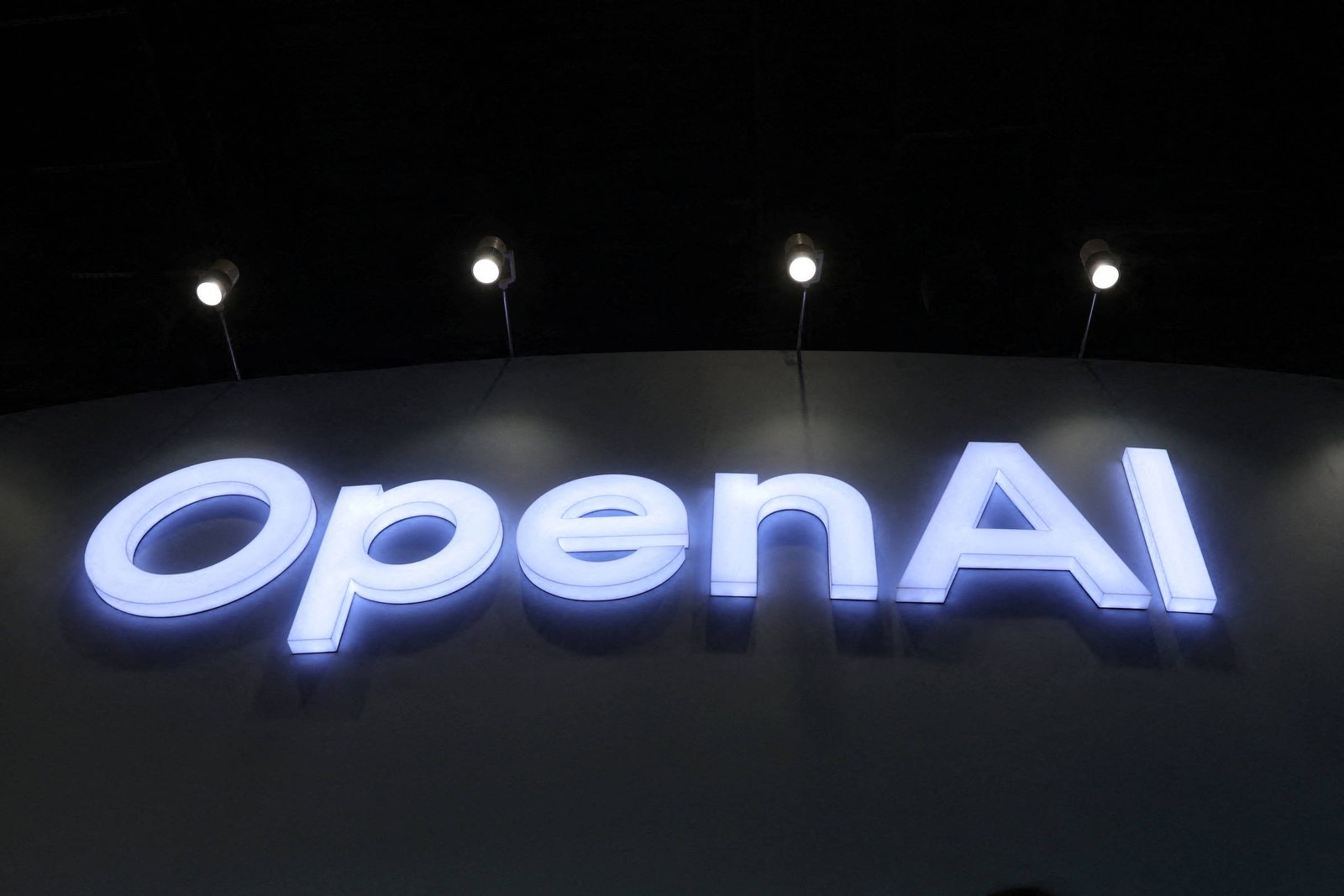Logo da OpenAI iluminado em branco contra um fundo escuro, com pessoas em sombra na parte inferior da imagem.